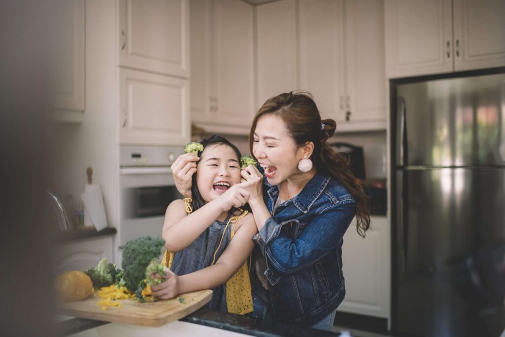 An Asian mother bonding with her daughter in the kitchen, playing with broccoli. 