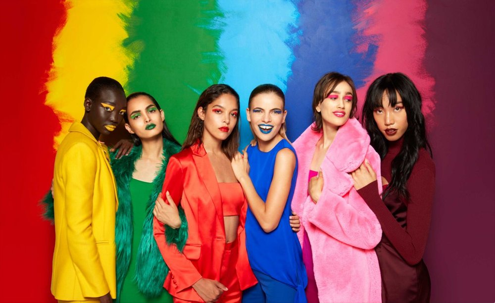 Models of various races stand in front of a rainbow colored wall. 