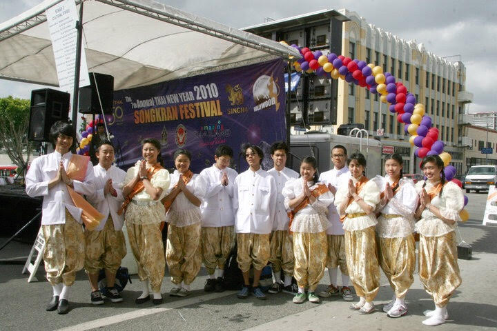 A large group of Thai American dancers in white shirts and gold patterned pants pose in front of a stage banner reading “Thai New Year 2010 Songkran Festival.” They are outdoors, standing under a canopy with colorful balloons decorating the area.