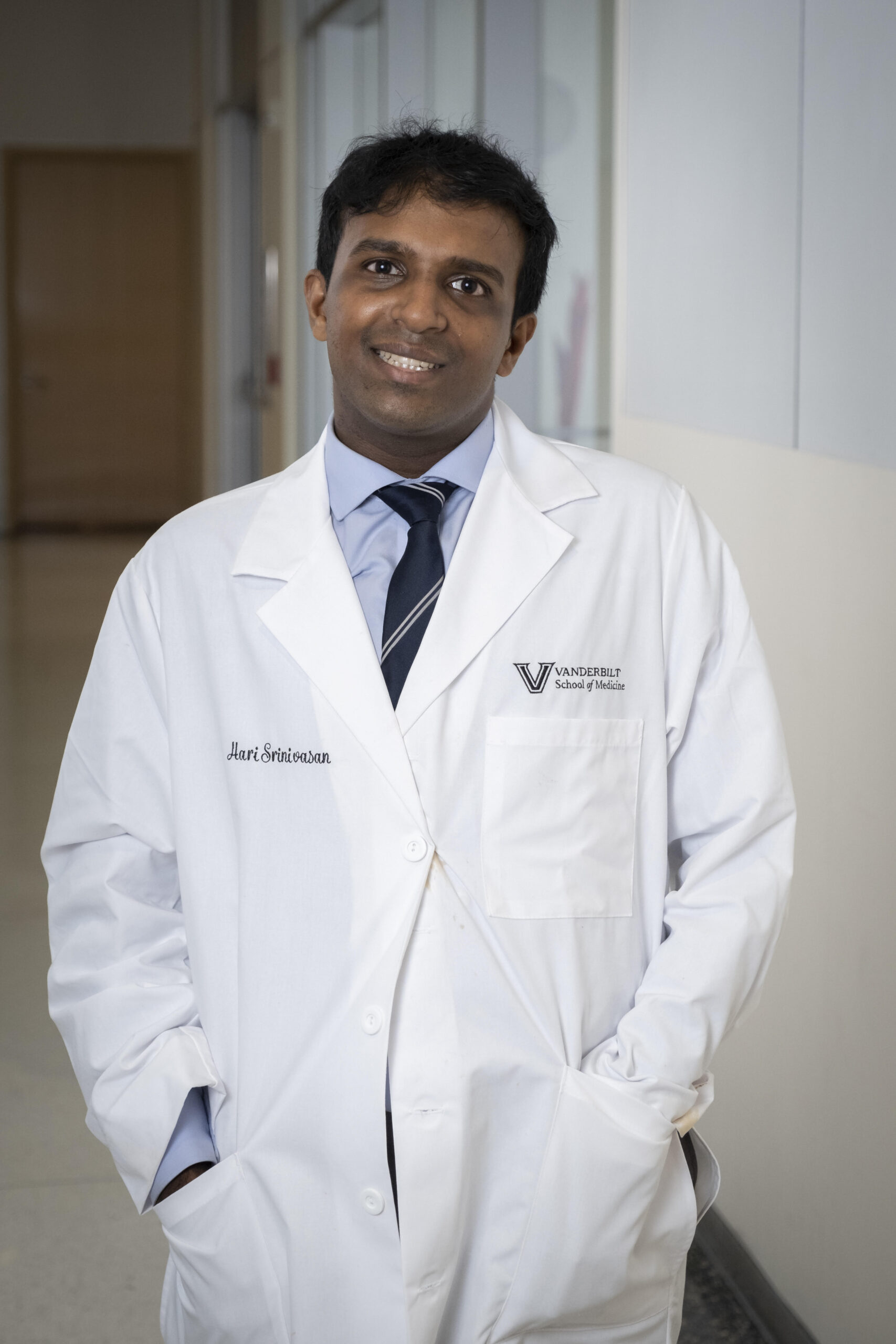 Hari Srinivasan, a minimally speaking autistic Indian American neuroscientist, poses in a hallway wearing a white lab coat embroidered with his name and "Vanderbilt School of Medicine." He smiles gently, with his hands in his coat pockets, dressed professionally in a shirt and tie.
