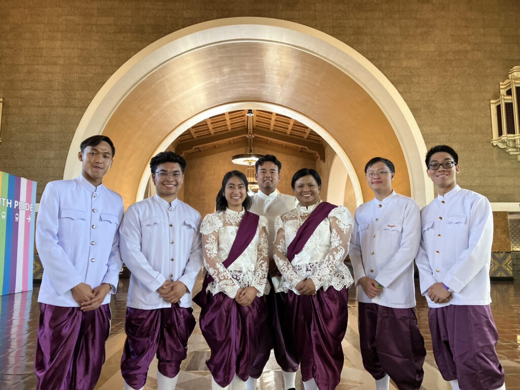 A group of seven people pose in traditional Thai clothing in front of a golden archway inside Union Station in Los Angeles. The women wear intricate white lace tops with maroon silk sampots, and the men wear white shirts with matching maroon pants. A “Youth Pride” banner is visible to the left.