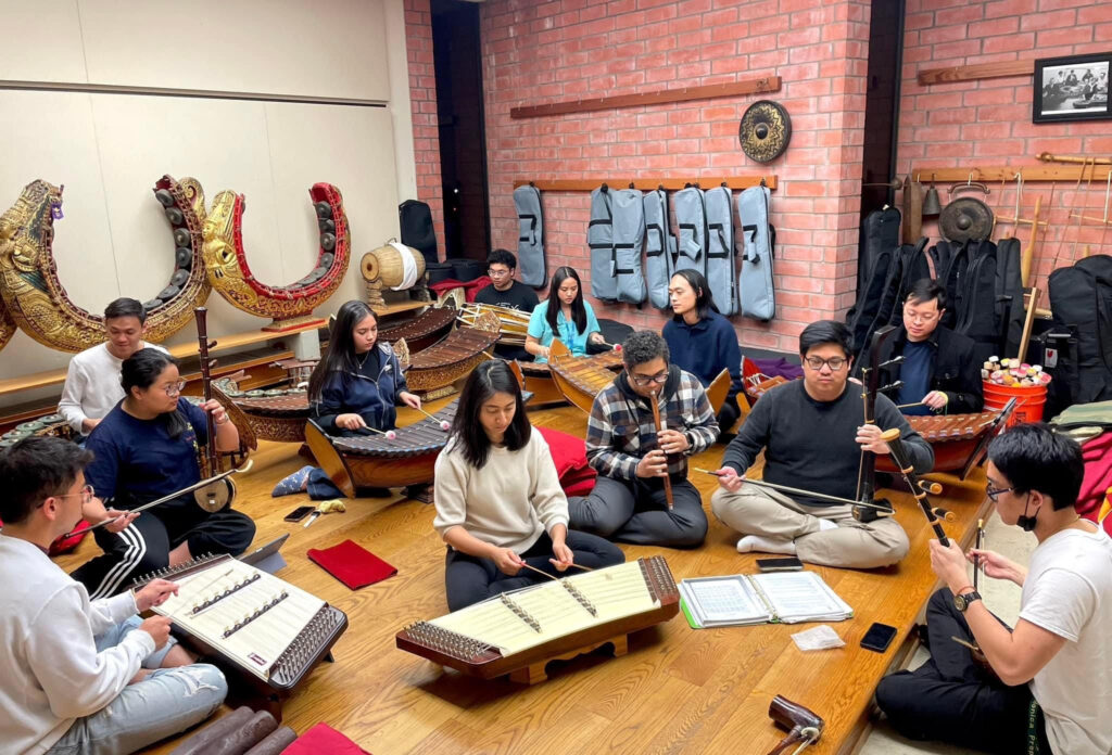 A diverse group of musicians, mostly seated on the floor, rehearse with traditional Southeast Asian instruments including khim (hammered dulcimer), saw (string instruments), and wooden flutes. They are surrounded by percussion instruments, gongs, and storage cases.