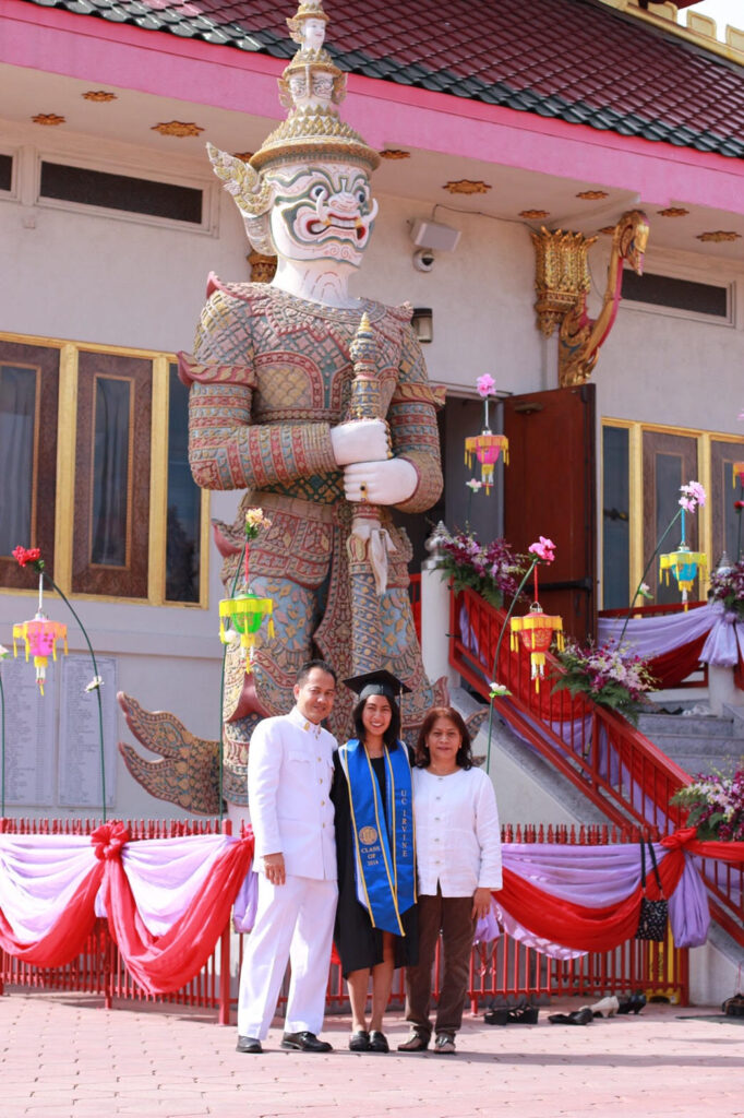 
A young woman in a graduation cap and gown poses smiling between two adults, likely her parents, in front of a large, ornately decorated Thai guardian statue (yaksha) at a temple. The graduate wears a blue sash that reads “UC Irvine” and “Class of 2014.” The family stands on a red brick courtyard adorned with pink, red, and purple fabric decorations and hanging paper lanterns. The temple staircase behind them is lined with floral arrangements, highlighting the celebration.