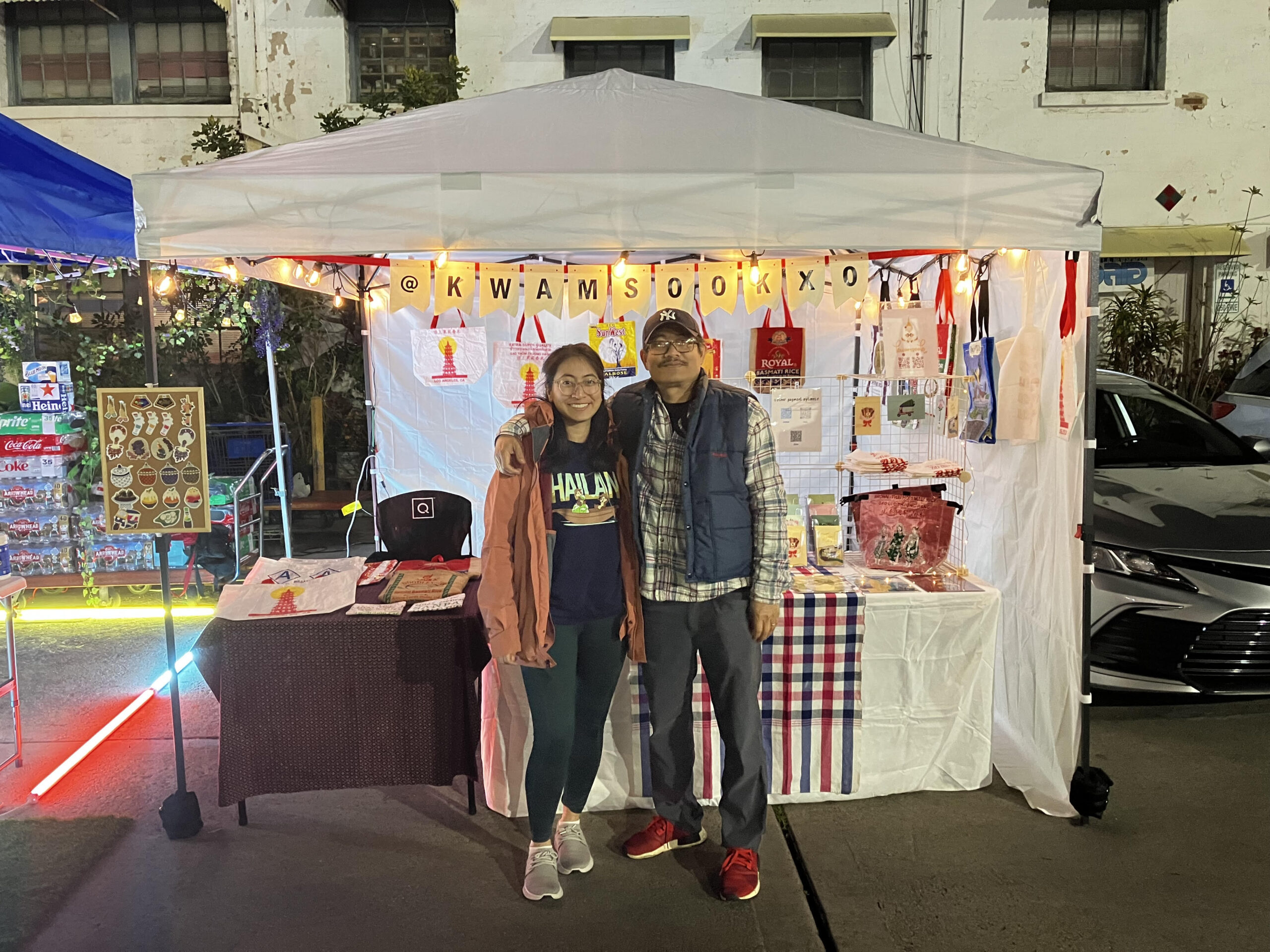 A smiling young woman and an older man stand arm in arm in front of a brightly lit vendor booth at a night market. The booth, decorated with string lights and traditional Thai items, displays textiles, aprons, and embroidered pieces. A sign above reads "@KWAMSOOKXOXO." The woman wears a coral jacket and dark pants, while the man is dressed in a plaid shirt, blue vest, and red shoes. Colorful products and crafts are neatly arranged on the tables inside the booth. The setting is outdoors, with neighboring booths and parked cars visible.