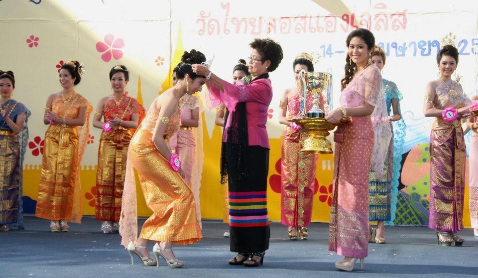 A pageant winner in a gold and orange Thai traditional dress bows as an older woman places a crown on her head during a Thai New Year ceremony. Other women in traditional dresses look on from the background as part of the celebration.