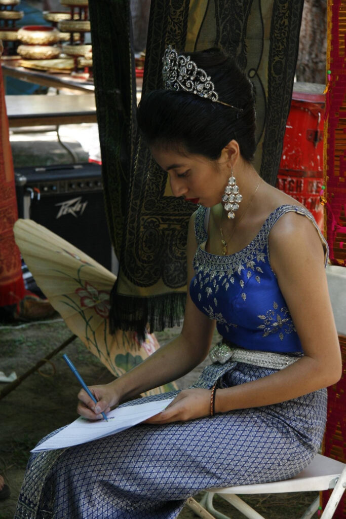 A woman wearing a blue traditional Thai dress with silver embroidery sits gracefully while writing on paper. She wears a silver tiara, chandelier earrings, and a belt, with traditional textiles and musical equipment in the background.