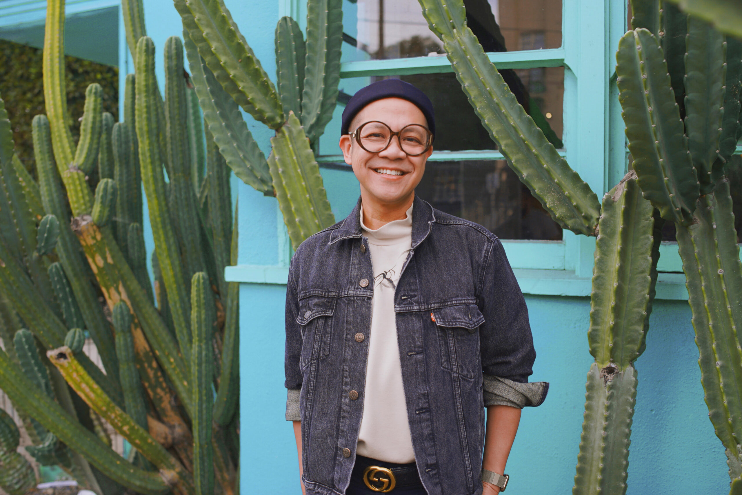 Jeremiah Abraham smiles brightly while standing outside against a turquoise building wall with tall green cacti. He wears a denim jacket over a light turtleneck, accessorized with a black Gucci belt, round glasses, and a navy beanie, giving a casual yet stylish vibe.