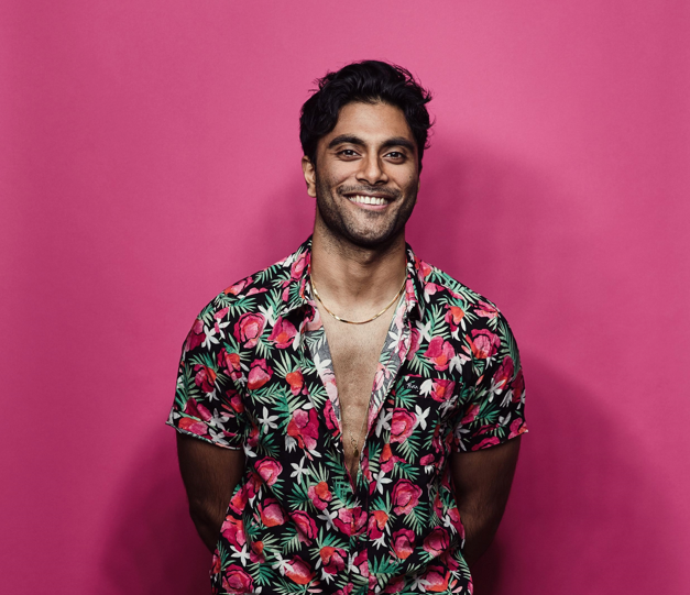 Akilla in a half buttoned floral Hawaiian shirt standing in front of a pink backdrop.