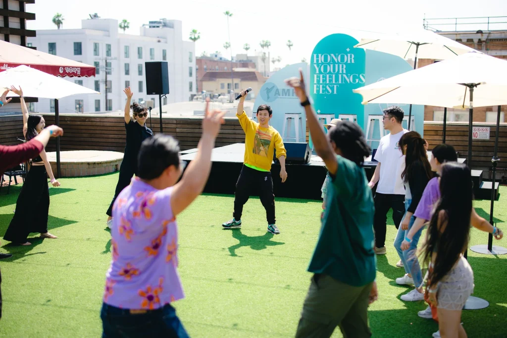 A diverse group of people joyfully participate in a dance or movement session on a sunny rooftop lawn during the "Honor Your Feelings Fest." A smiling instructor in a yellow sweatshirt holds a microphone and leads the group with an arm raised. Participants, wearing casual summer clothing, raise their hands and move energetically under large white umbrellas. A bright teal event backdrop and urban buildings are visible in the background, creating a vibrant, community-centered atmosphere.