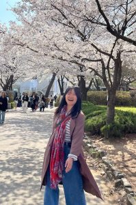 Ashley J. Chong is a Korean American woman with mid-length black hair, wearing a mauve jacket, striped shirt and red scarf. She is standing and smiling under cherry blossoms. 