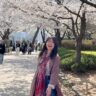 Ashley J. Chong is a Korean American woman with mid-length black hair, wearing a mauve jacket, striped shirt and red scarf. She is standing and smiling under cherry blossoms.