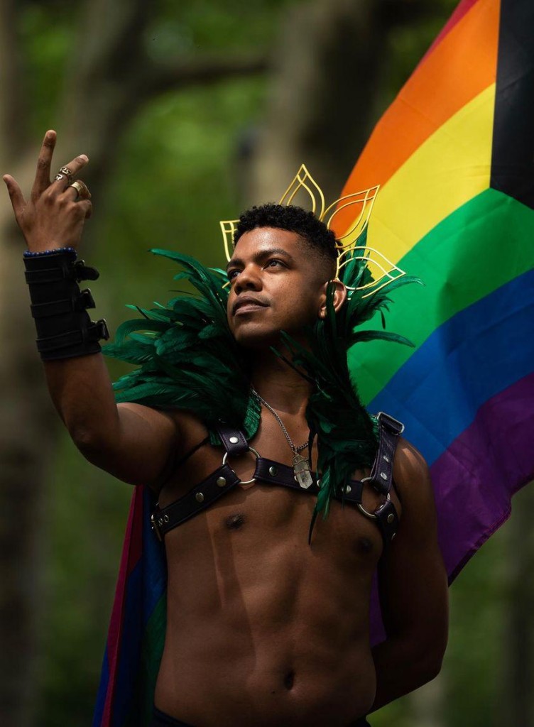 A person standing outdoors in front of blurred green trees holds up a peace sign with one hand. They wear a green feathered collar, a black leather harness, and a decorative gold headpiece. A rainbow Pride flag is draped behind them, creating a colorful backdrop. The person looks confidently into the distance.