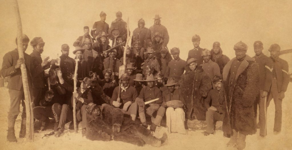 Sepia-toned historical photograph of a large group of African American soldiers posed outdoors in winter conditions. The men wear late 19th-century U.S. Army uniforms, some with fur coats, hats, and rifles. Several soldiers sit in the front row while others stand behind them in rows. Snow covers the ground, and the group faces the camera in a formal, posed arrangement.