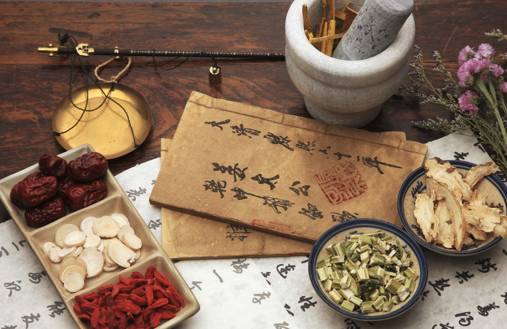 A traditional Chinese medicine setup is displayed on a wooden table, featuring dried herbs like red dates, goji berries, and sliced roots arranged in ceramic dishes. Handwritten scrolls with Chinese calligraphy lie beneath the herbs, alongside a stone mortar and pestle filled with dried ingredients. A brass balance scale and delicate flowers add to the cultural and ritualistic ambiance of the scene, evoking a sense of ancient healing practices.