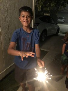 A young boy smiles while holding a lit sparkler outside at night. He wears a blue LA T-shirt and dark shorts, standing on a driveway near a parked car. The warm glow of the sparkler illuminates his face and hands.