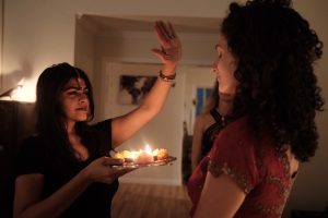 A woman in a black outfit holds a decorated plate with lit candles, performing an aarti ceremony for another woman wearing a red outfit. They are indoors, surrounded by soft, warm light and a calm atmosphere. The woman performing the aarti has a focused, serene expression.