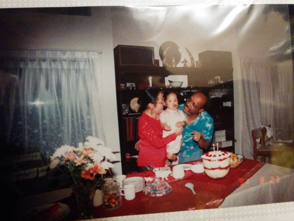 Printed family photograph of two adults holding a baby while standing behind a dining table with a birthday cake topped with lit candles. The adult on the left, wearing a red top, kisses the baby’s cheek, while the adult on the right, wearing a teal shirt, smiles and looks at the child. The baby wears a light-colored outfit. The table is covered with a red tablecloth and set with plates, cups, flowers, and a layered cake decorated with red accents. The scene appears to take place in a living room with curtains, a shelving unit, and warm indoor lighting, capturing an intimate birthday celebration.
