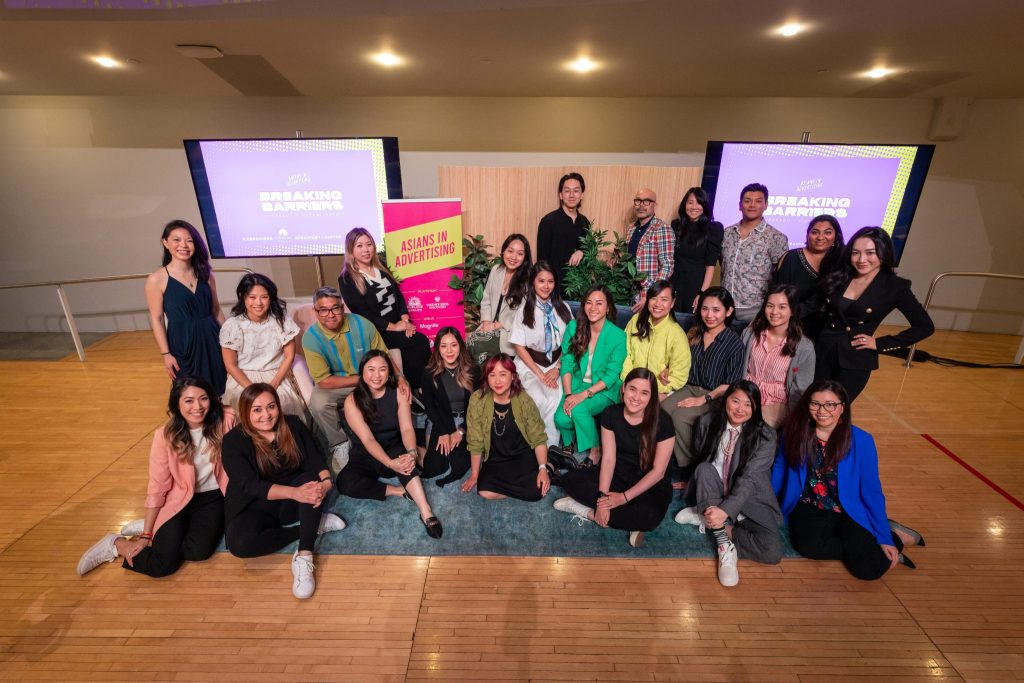 A large group of people pose together on a wooden stage floor for a group photo at a professional community event. Some are seated or kneeling in front while others stand behind. Two large screens in the background display event branding, and a vertical banner reads “Asians in Advertising.” The group smiles toward the camera in a celebratory, professional setting.