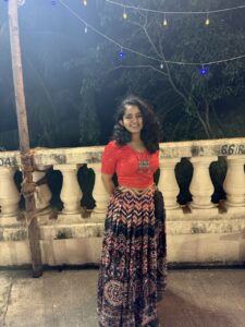 Prakruti smiles while standing outdoors at night in front of a white stone railing. She is wearing a red blouse with puffed sleeves and a long, patterned skirt in shades of pink, purple, and black. Colorful string lights hang above her, and trees are visible in the background under soft evening lighting.