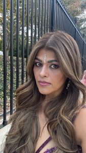 Falguni with long wavy hair and a jeweled bindi on her forehead poses outdoors in front of a black metal fence. She wears a magenta outfit with beadwork and matching earrings, and the background shows trees with autumn leaves.