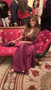 Falguni sits gracefully on a bright pink and gold ornate couch in a formal setting. She wears a flowing magenta gown with a beaded bodice and matching shawl, holding a black clutch. The room is filled with guests in traditional attire, and the carpet features floral designs in red and gold tones.