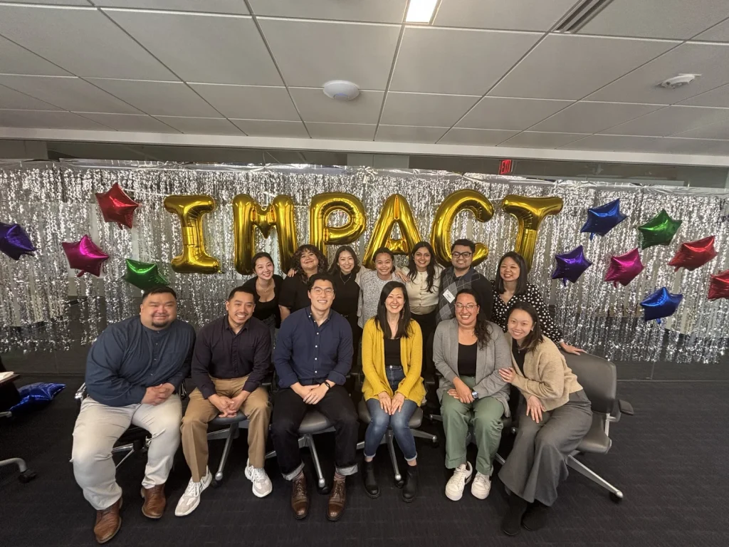 A group of about fifteen people pose together indoors for a cohort photo. They are arranged in two rows, with some seated on office chairs in front and others standing behind. A silver, sparkly backdrop with large gold balloon letters spelling “IMPACT” and colorful star-shaped balloons decorates the background. The group smiles toward the camera in a celebratory, professional setting, representing the LEAP Impact cohort.