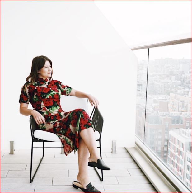 A woman sits in a black metal chair on a balcony overlooking a city skyline. She wears a fitted red dress with a bold floral pattern and black slip-on shoes. Her legs are crossed and one arm rests casually on the chair as she looks out through a large glass railing. The bright, minimal setting and soft daylight create a calm, reflective atmosphere.