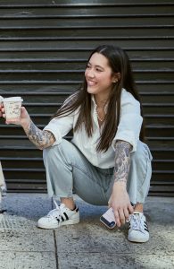 Meeja Kinsey is a mixed-race Korean American decendant of Korean adoptees (DoKAD). She has long brown hair, a fair complexion, tattooed arms and is squatting holding a coffee cup and smiling. 