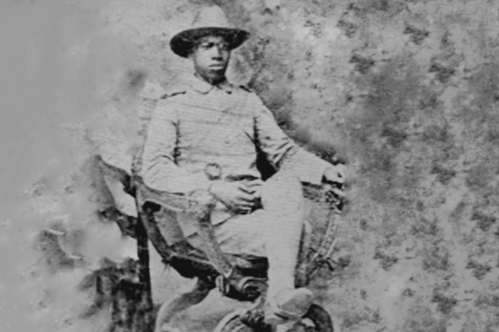 Black-and-white studio portrait of an African American soldier seated in a wooden chair. He wears a U.S. Army uniform with a wide-brimmed campaign hat and boots. A sword or saber rests at his side. The backdrop is a plain studio setting, and the soldier looks directly at the camera with a composed expression.