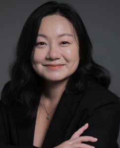 A professional studio portrait of Elaine Chiu with long, dark hair, wearing a black blazer and a delicate necklace. She faces the camera with a calm, confident smile against a dark, neutral background, her arms gently crossed, conveying warmth and professionalism.