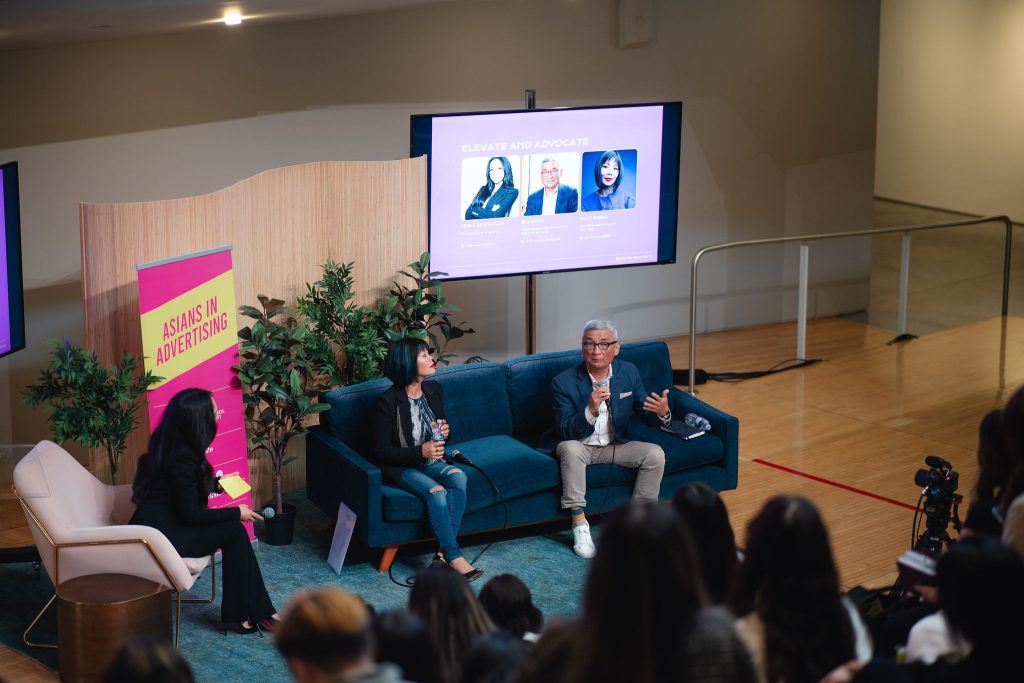 A panel discussion taking place on a small stage in front of an audience. Three panelists sit on modern couches holding microphones while speaking, with one person gesturing as they address the crowd. A screen behind them displays presentation slides with speaker headshots and the title “Elevate and Advocate,” and a vertical banner to the side reads “Asians in Advertising.” The setting appears to be a professional conference or summit focused on leadership and advocacy.