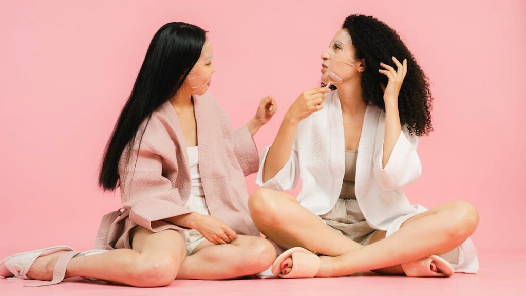 Two women sit cross-legged on the floor against a pastel pink background, enjoying a self-care session. Both wear lightweight robes and facial sheet masks. The woman on the left has long straight hair and a light pink robe, while the woman on the right has curly hair and wears a white robe, using a facial roller on her chin. They smile at each other, sharing a moment of relaxation and connection during a skincare ritual.
