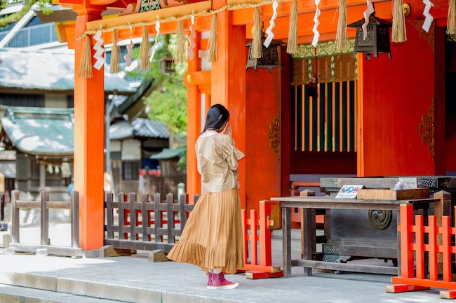 A woman stands in front of a brightly colored Shinto shrine in Japan, with her hands together in prayer. She wears a beige top, a flowing tan skirt, and red shoes. The shrine features vivid orange pillars, traditional wooden architecture, and hanging paper ornaments called shide. The peaceful setting is surrounded by trees and smaller temple structures in the background.