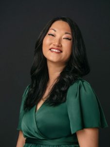 A professional studio portrait of Grace Young Foster with long, dark hair wearing a deep green dress. They smile warmly toward the camera against a dark, neutral background, with soft lighting that highlights their face and creates a polished, confident appearance.
