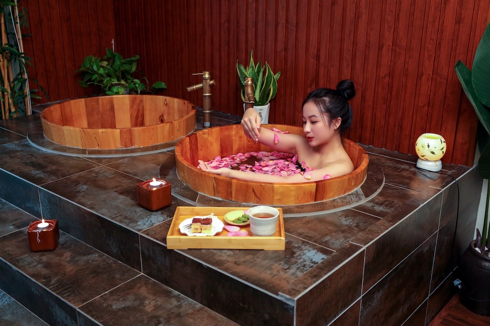 A woman relaxes in a wooden soaking tub filled with water and rose petals in a spa-like setting. She gently lifts a handful of petals while immersed in the warm bath. The room features dark tiled floors, wooden paneled walls, lush green plants, and soft ambient lighting from candles and a decorative lamp. A small tray with tea and traditional Asian sweets is placed on the edge of the tub platform, enhancing the atmosphere of tranquility and self-care.