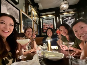 Five friends sit at a dimly lit restaurant table holding cocktails and smiling at the camera. Framed art lines the walls behind them, and a golden lamp glows on the table, casting warm light over their glasses and plates.