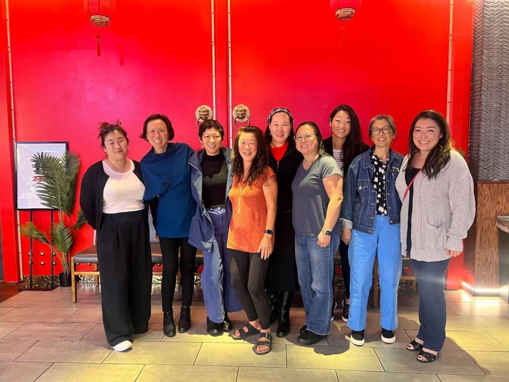A group of nine people stand together indoors in front of a bright red wall with gold lantern decorations. They smile toward the camera, with arms around each other, creating a sense of unity and celebration.