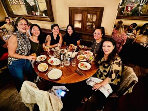 Seven people gather around a circular table at a restaurant, sharing a meal and smiling brightly. Plates, drinks, and food are spread across the table. The group poses together, creating a sense of warmth, friendship, and community.