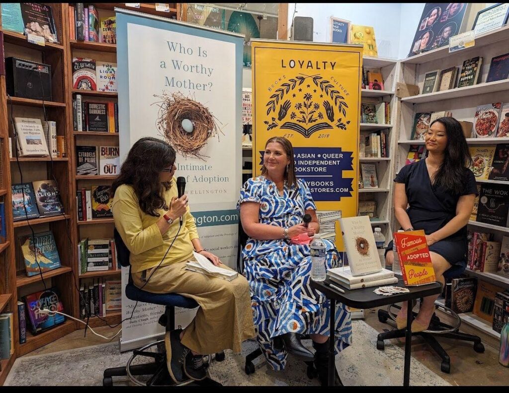Three people sit in conversation at a bookstore event surrounded by bookshelves. A banner behind them reads “Who Is a Worthy Mother? An Intimate History of Adoption.” Another sign reads “Loyalty Books – Asian, Queer, and Independent Bookstore.” The speakers are engaged in discussion, one holding a microphone and smiling.