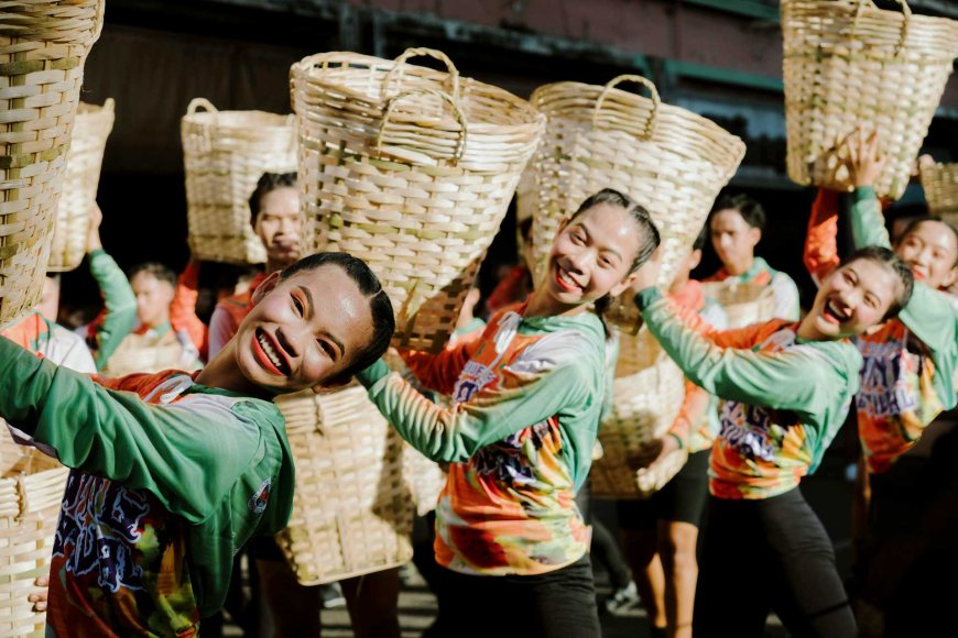 A group of smiling young Filipino performers in brightly colored costumes featuring green sleeves and tie-dye patterned tops joyfully dance in a street parade. Each person holds a large woven basket above their shoulder in a dynamic pose, forming a vibrant and synchronized display. The scene is full of energy and celebration, capturing the spirit of a traditional Filipino cultural festival.