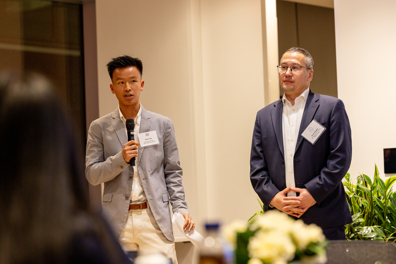 Two people stand at the front of a professional event space. One person on the left speaks into a handheld microphone while holding notes, wearing a light-colored blazer and white pants. Another person stands beside them with hands clasped, wearing a dark blazer and glasses. Both face an audience in a well-lit indoor setting.