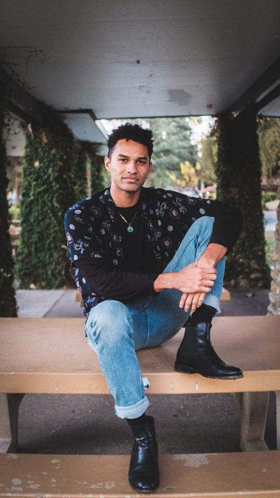 A person with short curly hair sits on concrete steps beneath a covered walkway lined with ivy-covered columns. They wear a patterned short-sleeve button-up over a black long-sleeve shirt, light blue cuffed jeans, and black leather boots. A green pendant necklace rests at their chest. They sit with one knee raised and arms resting casually, looking directly at the camera with a calm, confident expression.
